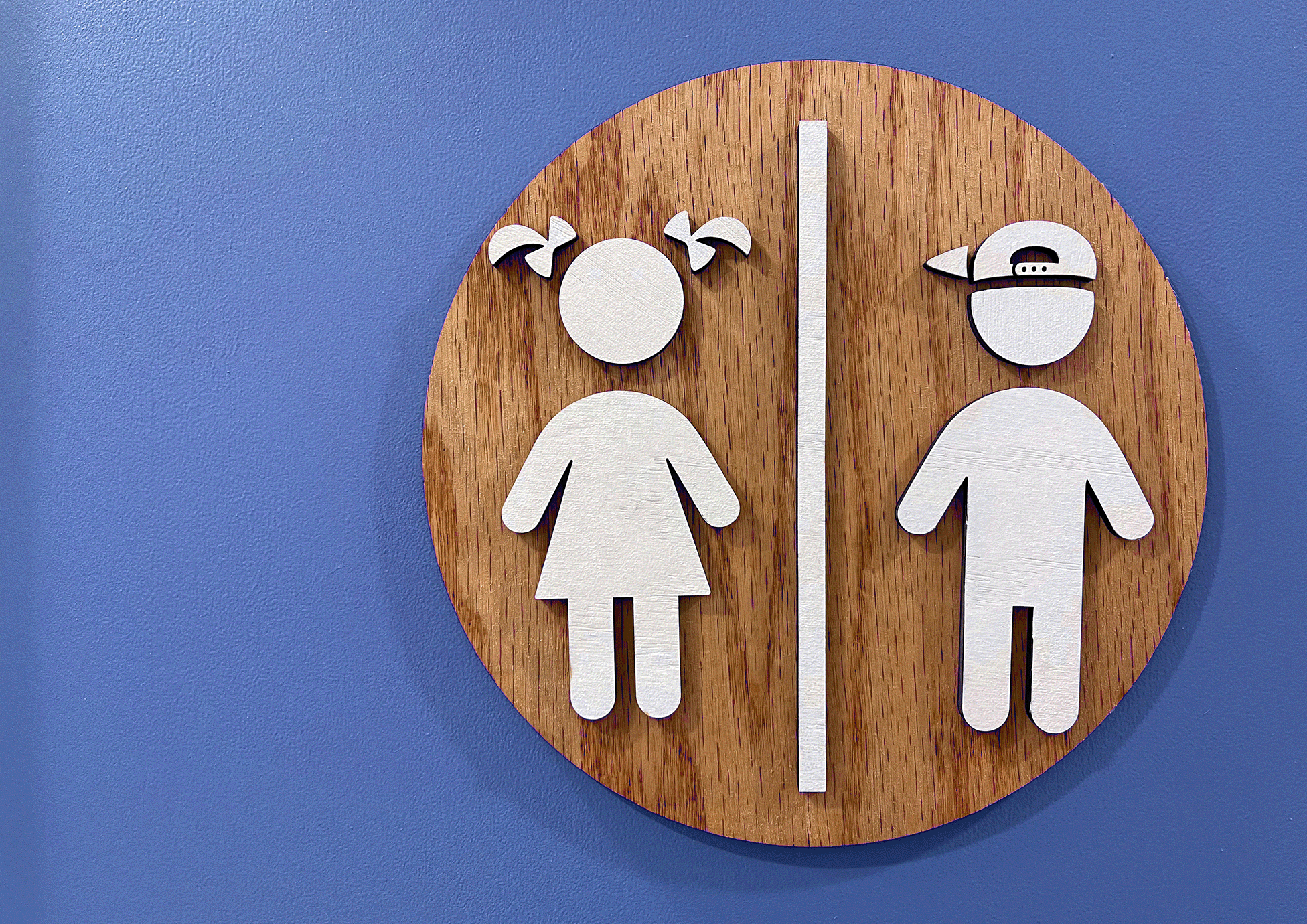 Rundes Toilettenschild aus Holz an einer blauen Wand. Auf dem Schild sind weiße Symbolfiguren für einen Jungen mit umgedrehter Basecap und ein Mädchen mit Zöpfen zu sehen, getrennt durch eine weiße Linie.